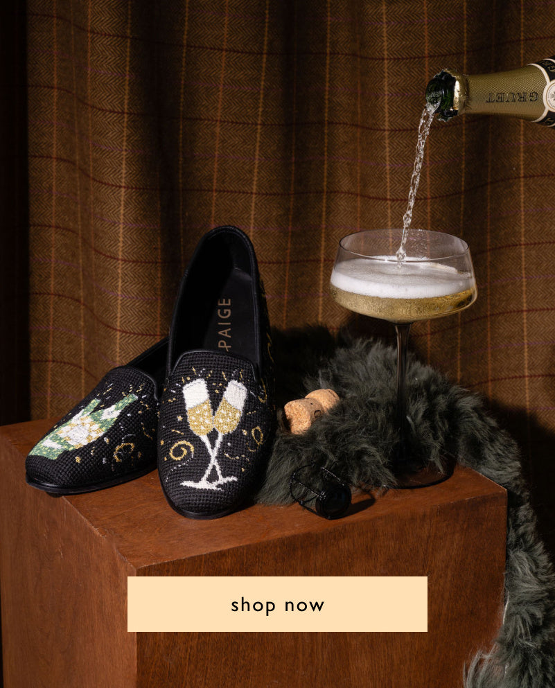 A pair of black handstitched needlepoint loafers with a champagne design, displayed alongside a needlepoint backdrop featuring a champagne motif. The loafers are adorned with text and decorative elements related to celebration and champagne.


