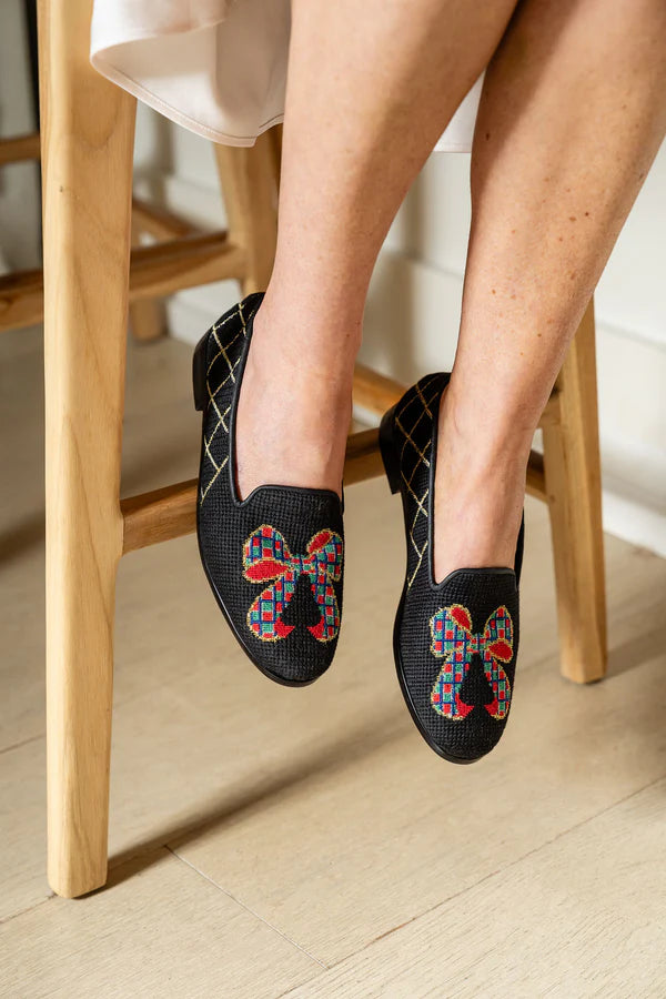 A pair of needlepoint loafers with a tartan bow design, featuring a black base with red, green, navy, gold, and silver accents,