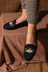 A black hand-stitched needlepoint loafer with a bee on a snaffle bit, worn by a person seated with plaid patterned legs.