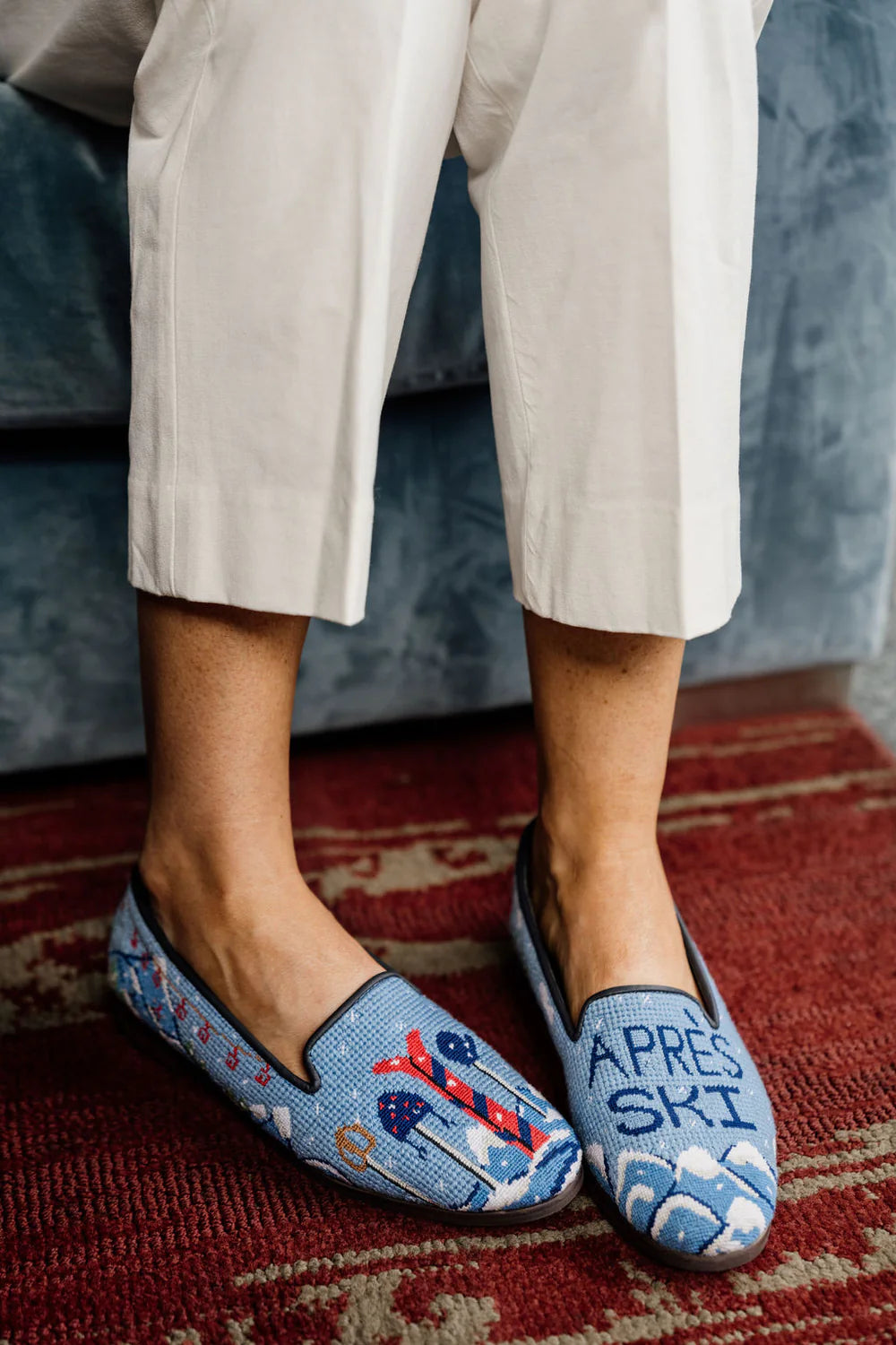 A pair of handstitched needlepoint loafers with a ski and mountain theme, featuring a blue backdrop and details of ski poles, skis, and 'Apres Ski' text.