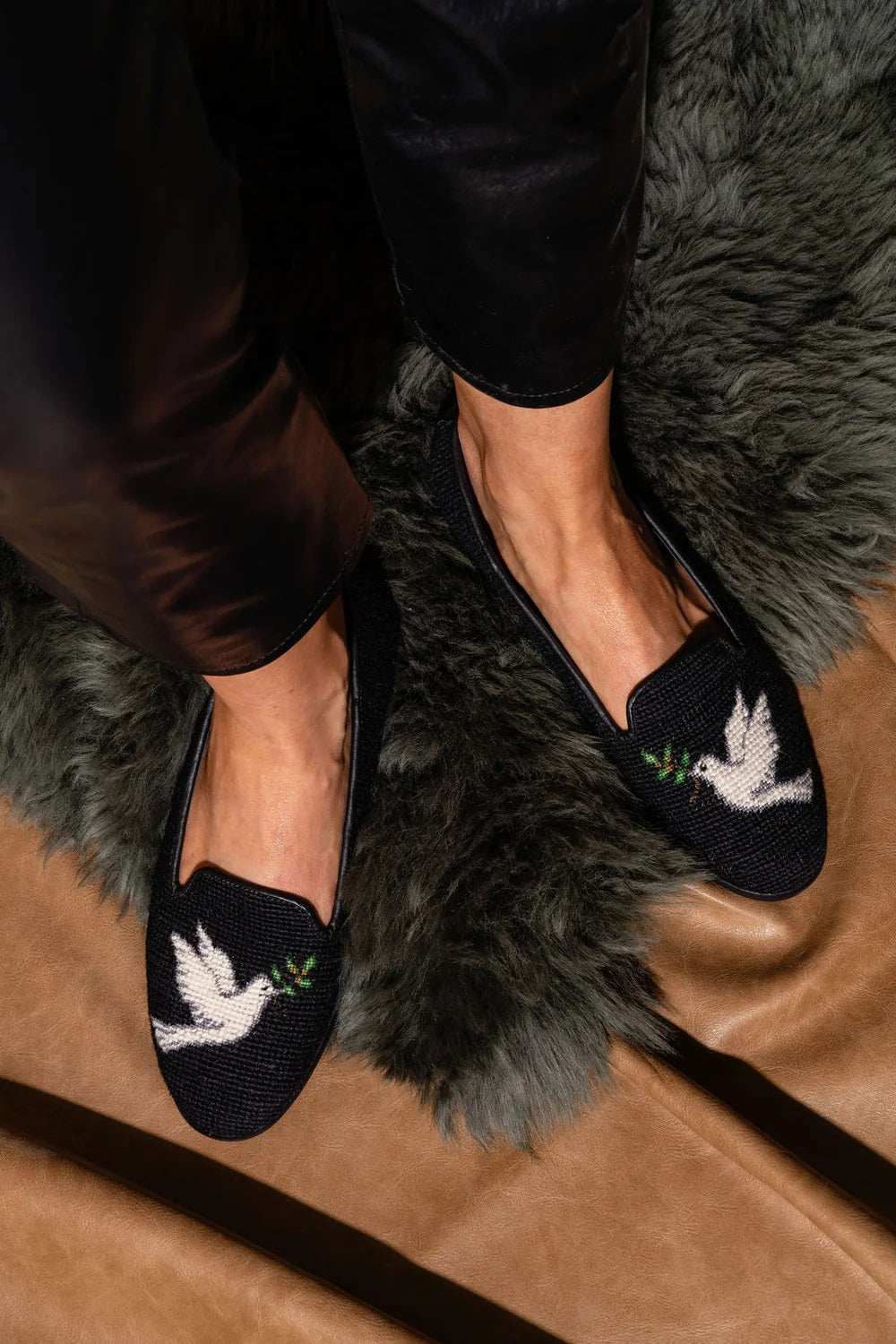 A pair of black needlepoint flats with a white dove design on the top, worn by a person with a tan background.