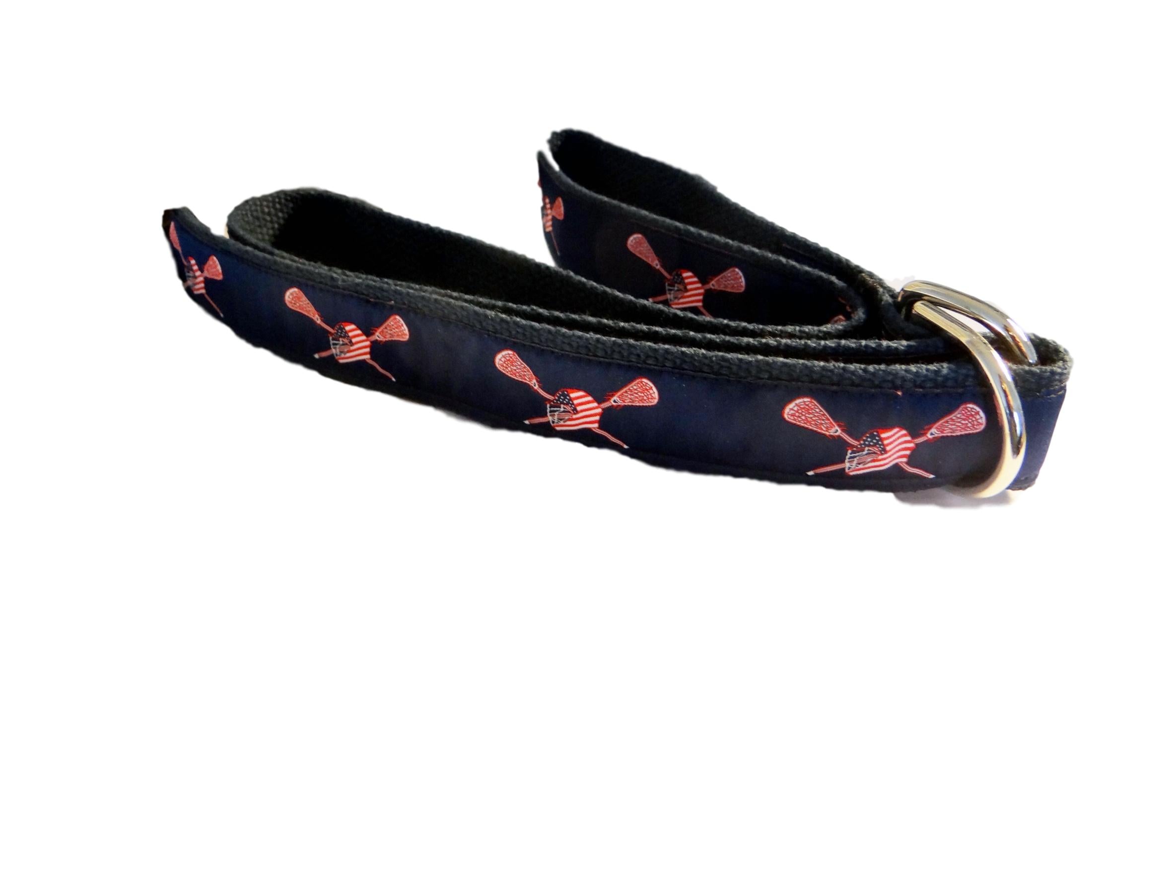 Custom, Hand stitched, Unisex Lacrosse D-Ring Ribbon Belt is a fav with players and coaches. Its features are drawn on a navy ribbon background with red and white sticks and a red white and blue helmet as the primary feature. It is finished  on navy webbing with stainless D-Rings.